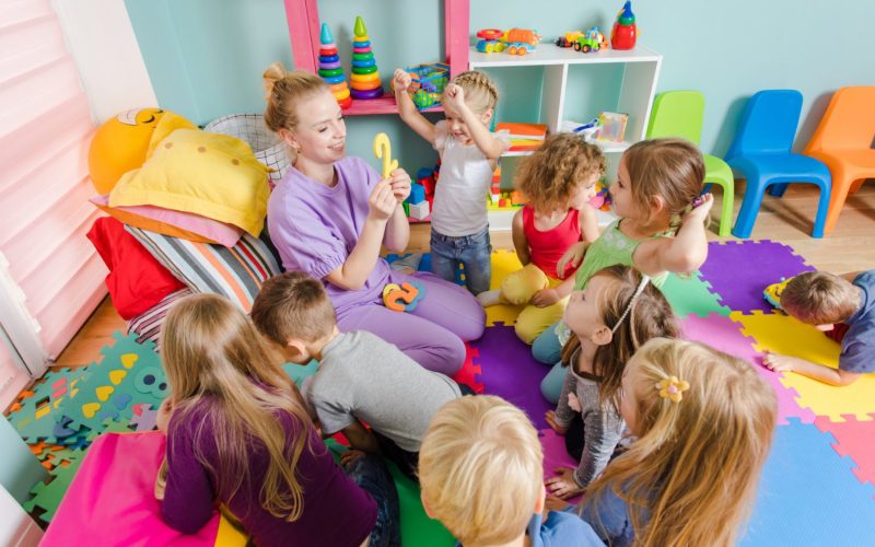 Young engaged teacher sitting with group of preschool children on a floor during the lesson. Process of learning can be fun. Room with colorful mats on a floor and multicolor furniture.