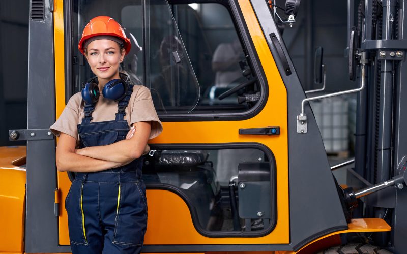 Attractive female standing next to forklift truck driver in an industrial area in factory, looking at camera confidently, dressed in uniform. good job. female engineer at work place